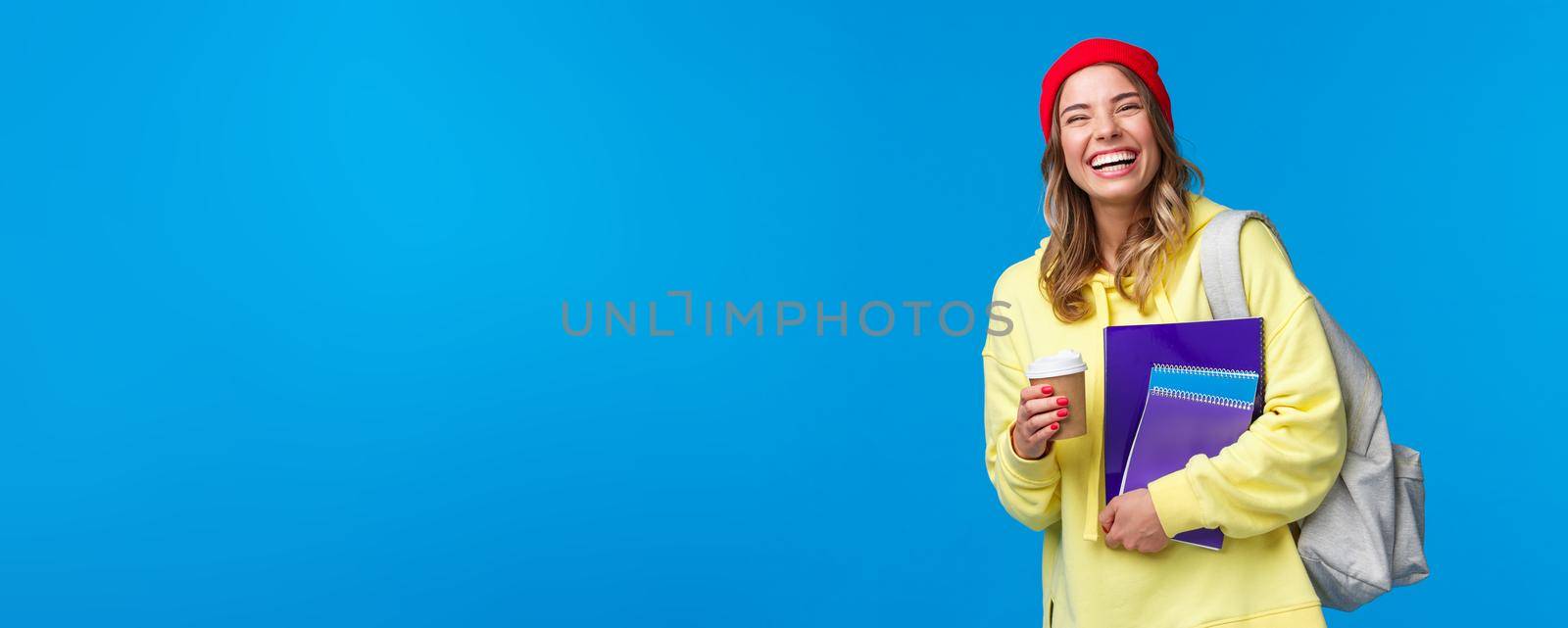 Cheerful happy female student having fun, enjoying university lifestyle, carry backpack and notebooks, drinking take-away coffee, talking to classmates, standing blue background joyful by Benzoix