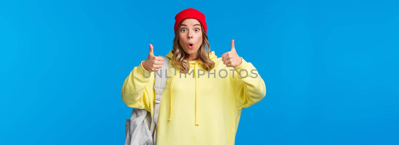 Wow really cool, good job. Impressed good-looking blond young female student in red beanie and yellow hoodie, carry backpack, praise friend nice work, show thumbs-up in approval or like.