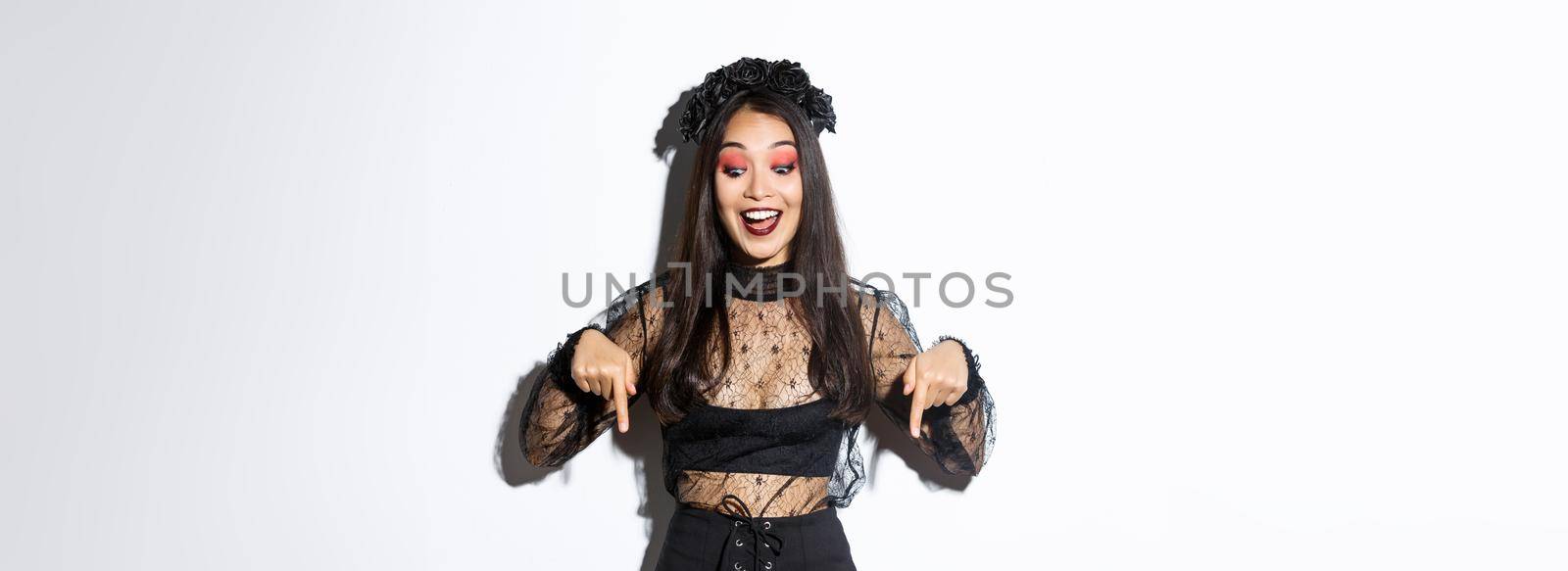 Excited asian woman dressed-up for halloween, smiling amused and pointing fingers down, showing advertisement, standing in party costume over white background.