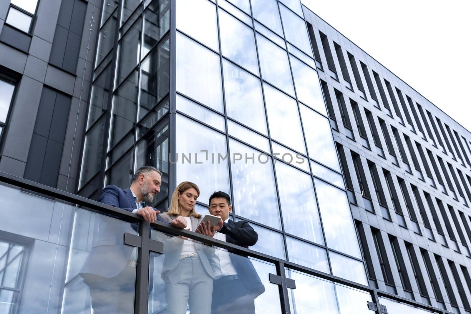 Three business people woman and man, outside office building, serious and thoughtful discussing and looking at electronic tablet, Asian man and woman in business suits