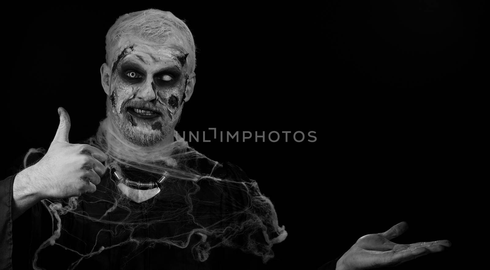 Creepy man with bloody scars face, Halloween stylish zombie make-up. Scary wounded undead guy showing thumbs up and pointing at left on blank space. Place for your advertisement logo. Sinister monster