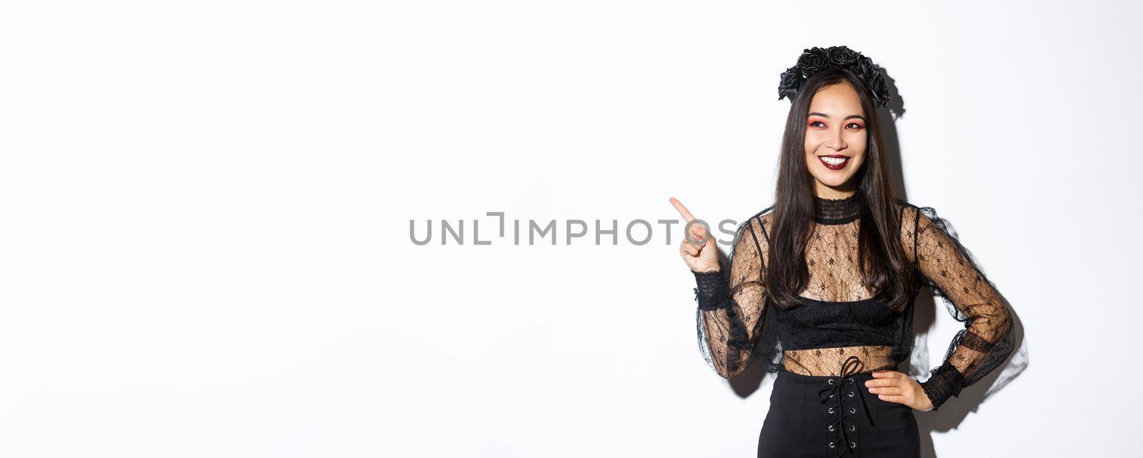 Satisfied smiling asian woman in evil witch or banshee costume celebrating halloween, looking pleased and pointing finger upper left corner, showing your promo banner, white background.