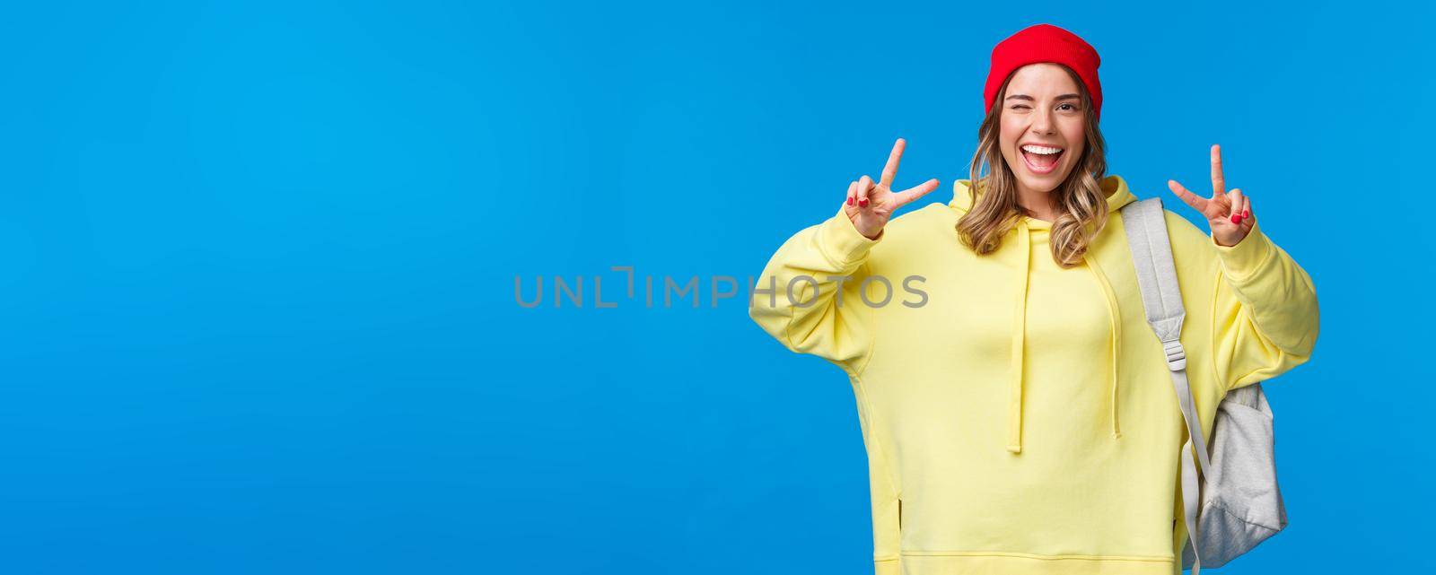 Back to school concept. Cheerful hipster girl with blond hair in beanie, carry backpack, wink carefree and smiling, showing peace gesture send positive vibes, stand blue background.