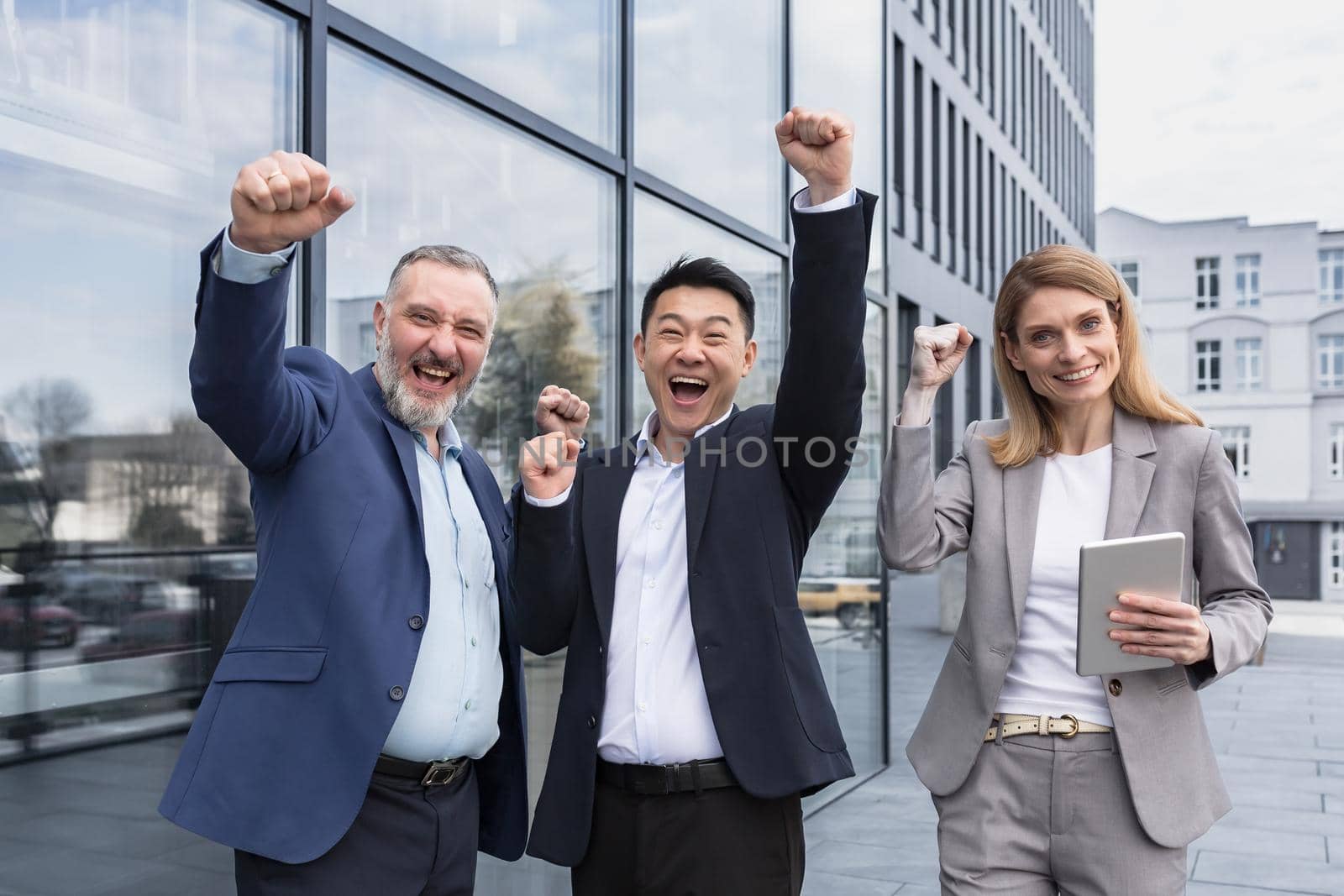 Successful diverse business team, three loving men and woman looking at camera and happy celebrating victory, team dreaming outside office building, business group in business suits