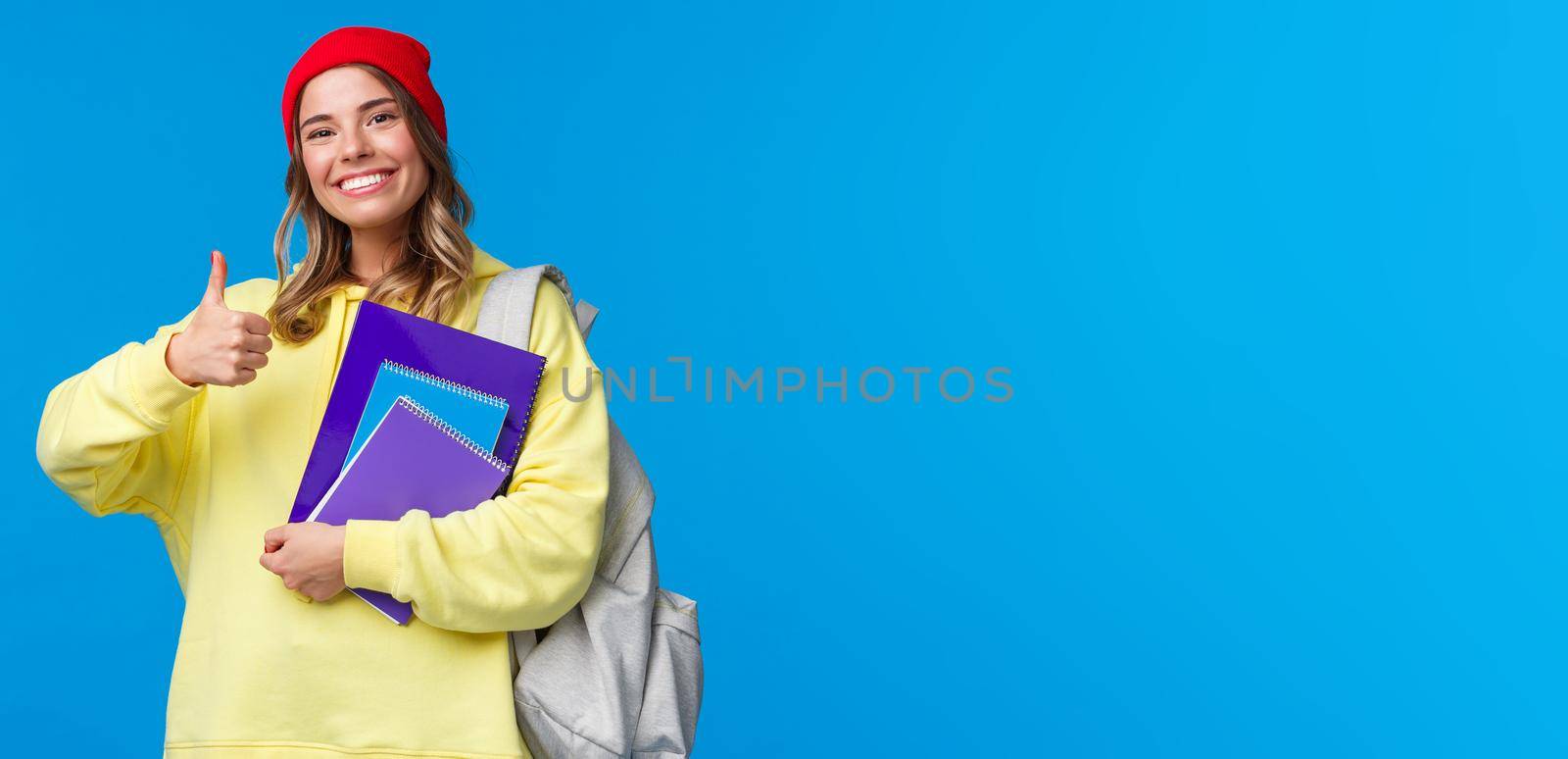 Sounds good. Cheerful cute and smart caucasian girl in red beanie studying hard for exams, encourage student keep up, show thumbs-up in like or approval, hold notebook and backpack.