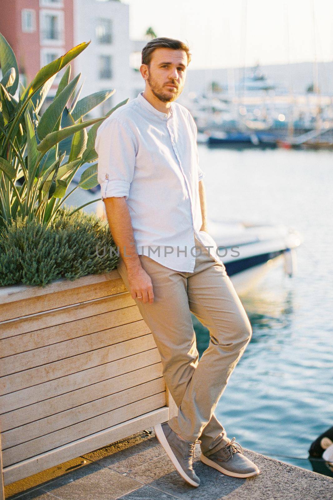 Young man stands on a pier leaning against a wooden tub with green plants. High quality photo