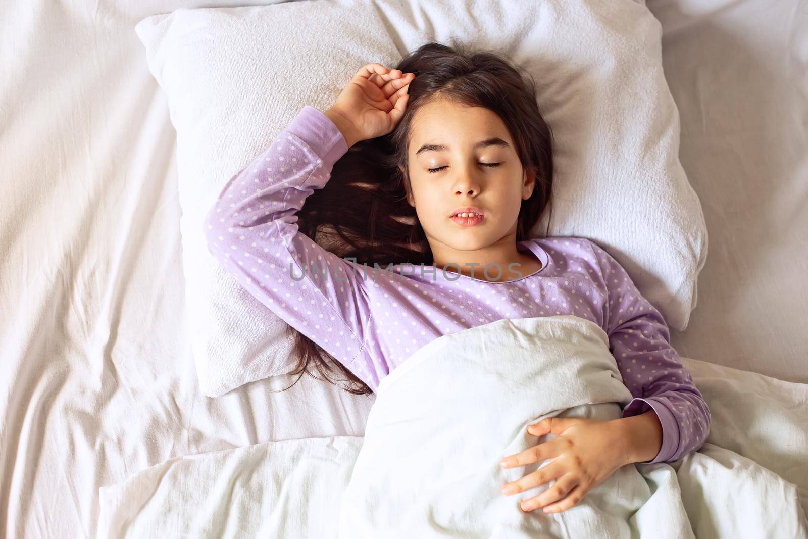 Little brunette girl in purple pajamas, sleeps lying on a white pillow on the bed. View from above. Top view. Copy space