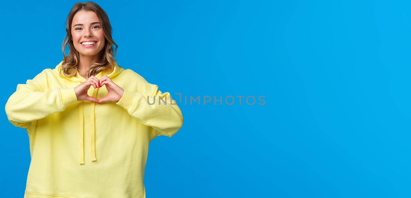 Lovely caring cute blonde girl in yellow hoodie showing heart gesture and smiling as express symapthy, confess in love or heartwarming feelings towards person, stand blue background.