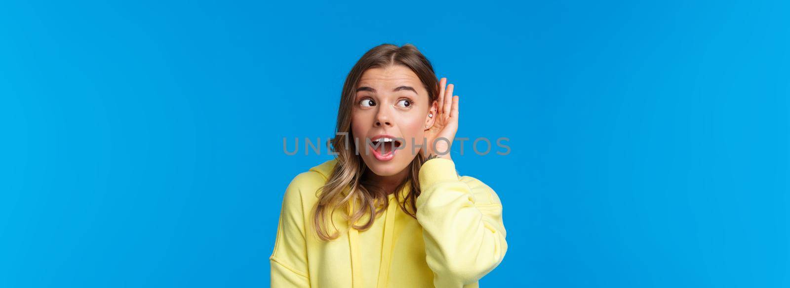 Close-up portrait of intrigued and curious blond young girl with pierced ear, gossiping, eavesdropping and listening to someone conversation interested, standing blue background. Copy space