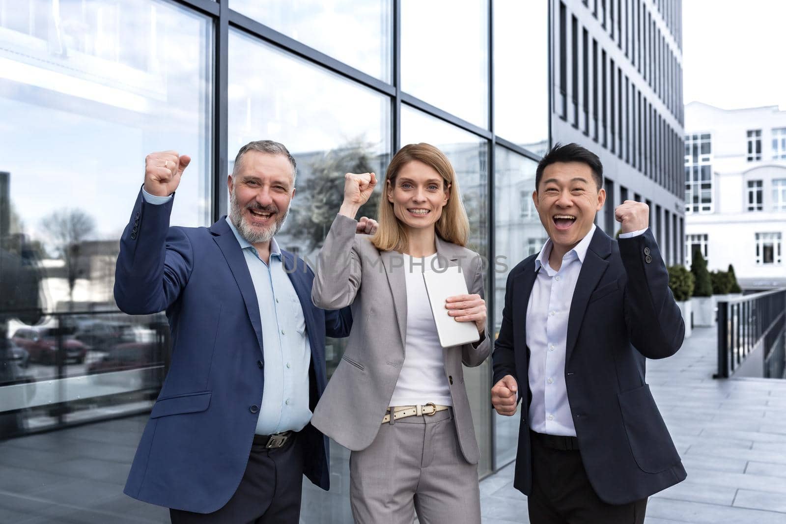 Successful diverse business team, three loving men and woman looking at camera and happy celebrating victory, team dreaming outside office building, business group in business suits