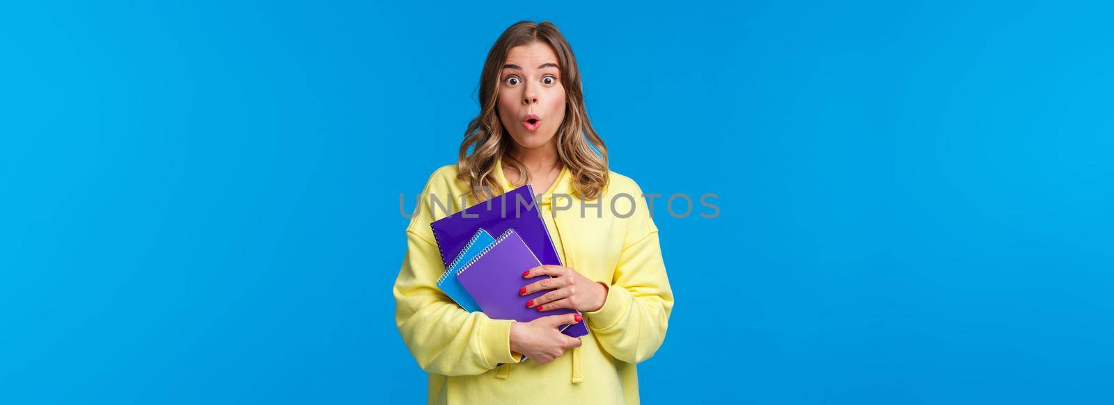 Waist-up portrait of surprised and wondered blonde caucasian female say wow and express amazement hear interesting news, gossiping in college, holding notebooks and essay, blue background.
