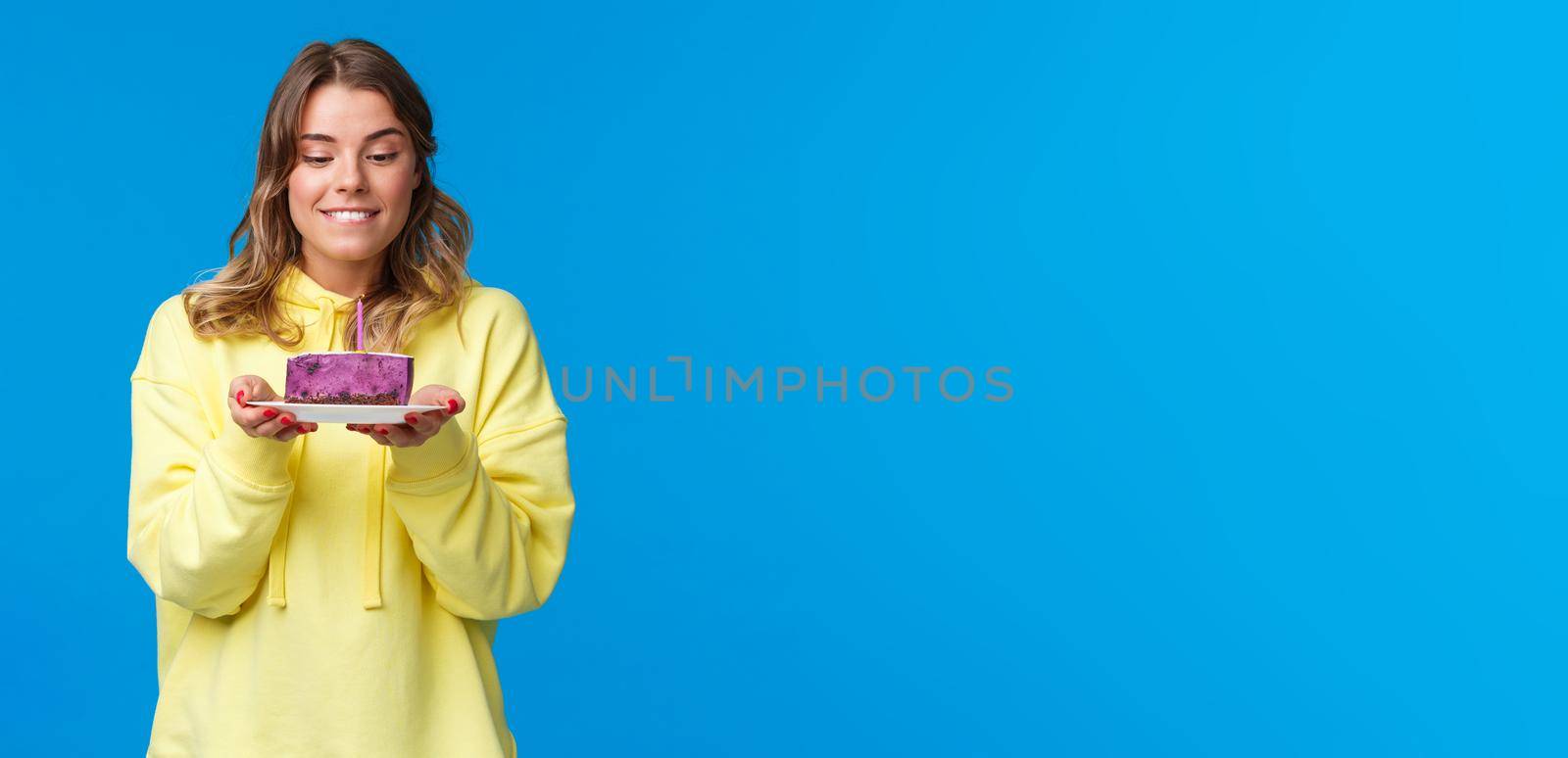 Celebration, party and lifestyle concept. Girl want have bite of tasty desert, holding b-day cake with lit candle as making wish, look with desire and happiness, celebrating birthday, blue background.