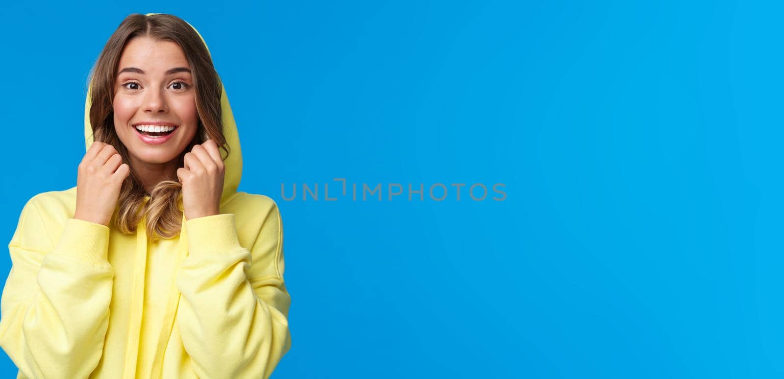 Lifestyle, people and youth concept. Close-up portrait of beautiful smiling blond female put on hoodie, looking camera with upbeat enthusiastic expression, standing blue background.