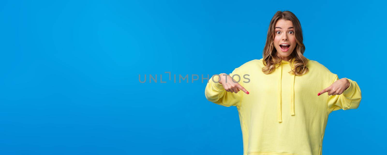 Wow thats something new. Impressed good-looking blond girl telling about awesome promo offer, best discounts, pointing fingers down and looking camera as suggest buy it, blue background.