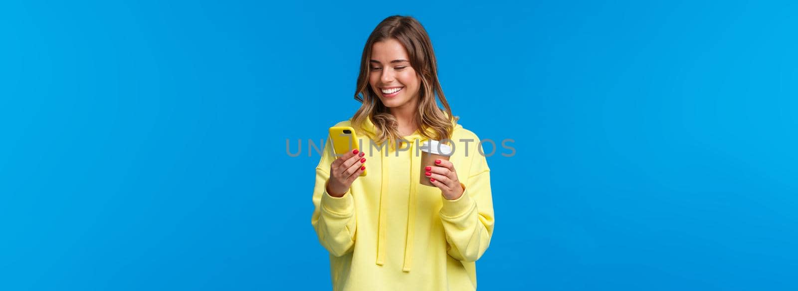 Carefree joyful european female in yellow hoodie, holding take-away coffee and browsing internet in mobile phone, texting friend laughing over funny video or meme, blue background by Benzoix