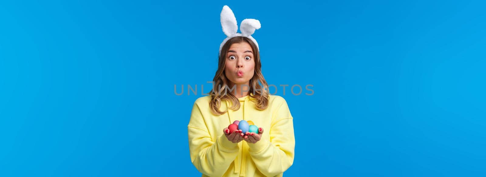 Holidays, traditions and celebration concept. Silly cute caucasian blond girl present you Easter eggs, painted it for holiday, show mwah kiss expression and wear rabbit ears, blue background.