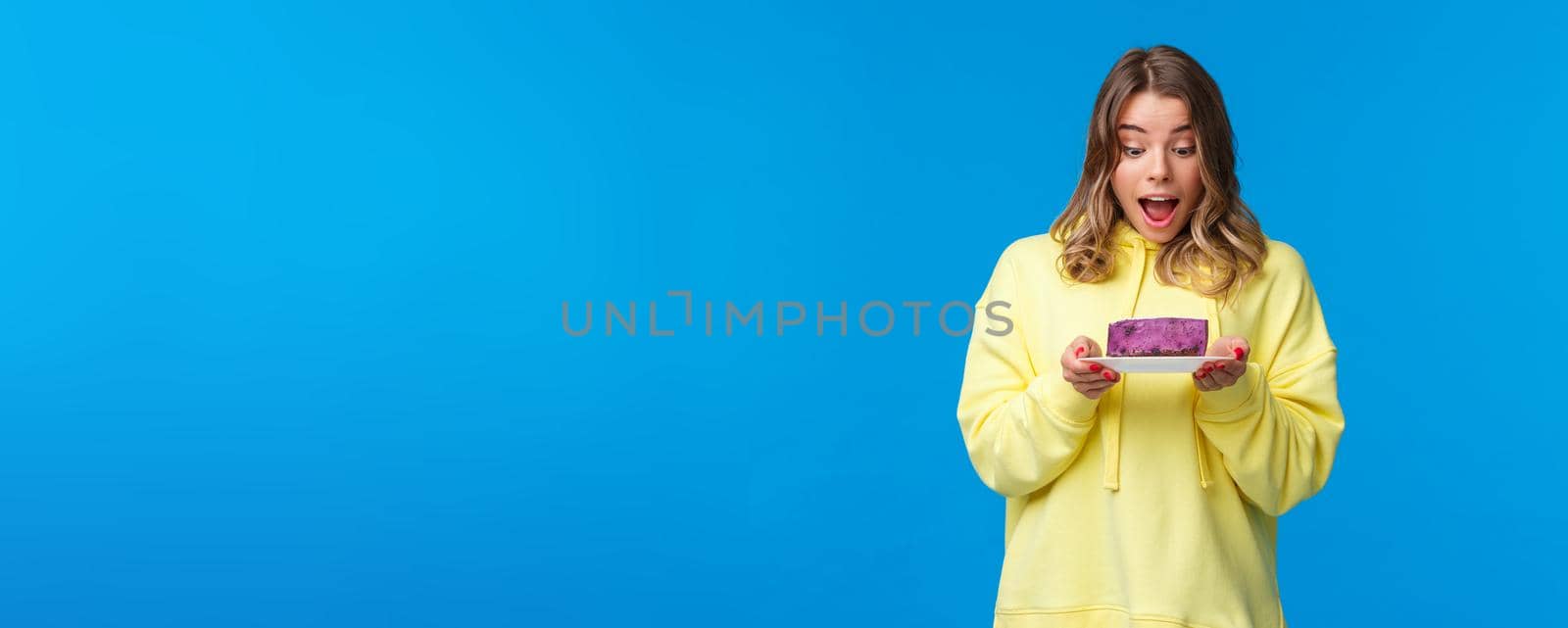 Celebration, party and lifestyle concept. Happy and pleased blond caucasian girl in yellow hoodie, looking fascinated at tasty berry cake with desire, want have bite, stand blue background.