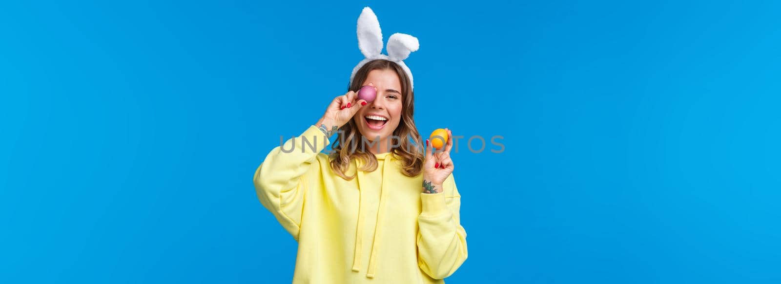 Holidays, traditions and celebration concept. Happy cheerful young pretty female celebrating Easter day, showing two painted eggs and laughing, wearing cute rabbit ears, blue background.
