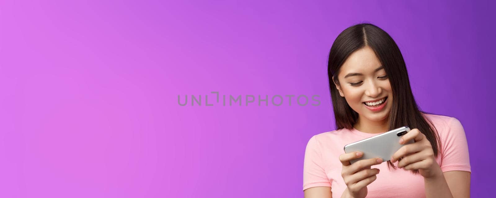 Close-up joyful attractive asian woman brunette having fun spend time playing smartphone game, laughing smiling eager win race, hold phone horizontal beating score, purple background.