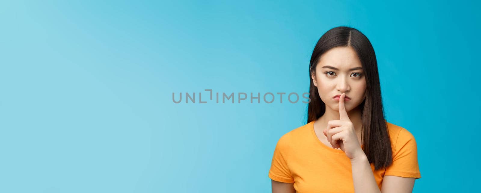 Hush stay silent. Serious-looking asian girl look displeased grumpy show shush sign press index finger lips, frowning upset, telling not speak, untell secret, prohibit loud music, blue background.