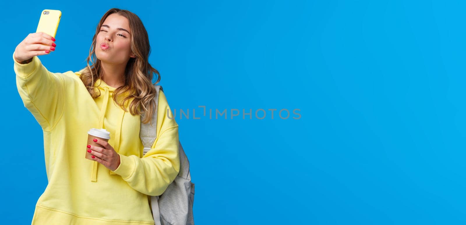 Cute positive and feminine beautiful young female student heading college holding paper cup from favorite cafe and backpack with books, taking selfie show kiss mwah face at mobile camera.