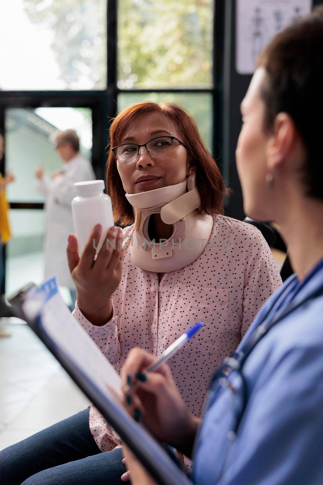 Woman with cervical neck collar holding pills bottle while nurse writing medication treatment on report during examination. Injured patient having appointment in hospital waiting area