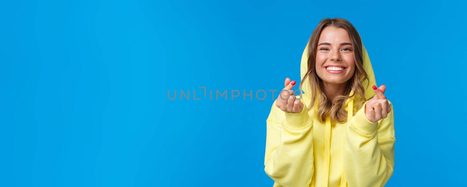 Close-up portrait of cute and silly blond caucasian girl in yellow hoodie, showing korean heart gesture and smiling, vibing, enjoying party, having fun over blue background. Copy space