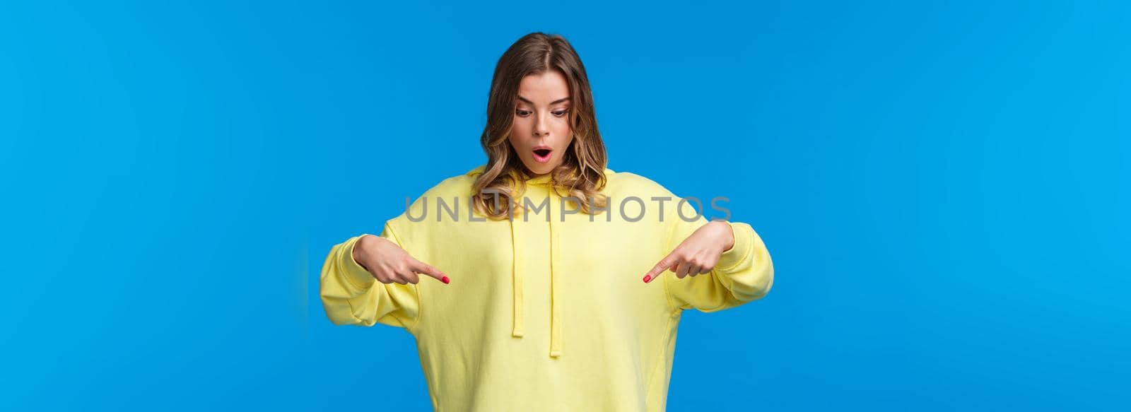 Girl found something stunning. Amused and speechless surprised blond alluring woman in yellow hoodie, drop jaw stare and pointing fingers down at cool product, blue background.