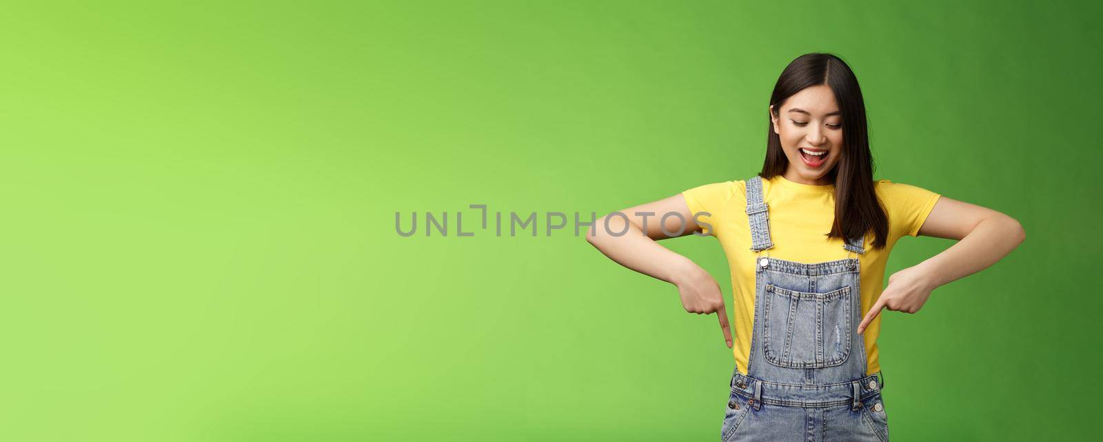 Impressed entertained cute asian girl watching pointing down interested, observe cool promo, smiling broadly, fascinated gaze bottom advertisement, take look curious thing, green background.