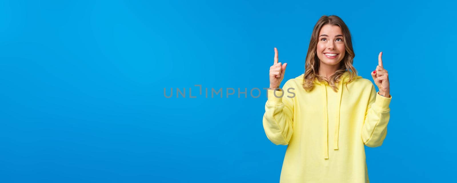 Happy alluring caucasian female with blond hair in yellow hoodie, searching career opportunities, found something interesting, looking and pointing fingers up, smiling pleased.