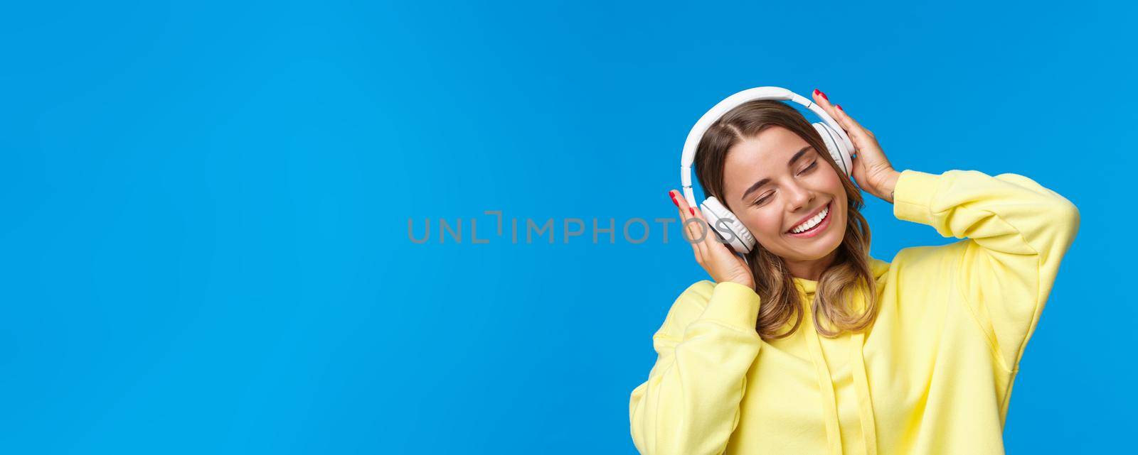 Music, lifestyle and youth concept. Close-up portrait of joyful cute young blond girl listening to favorite song in headphones, vibing, smiling delighted, standing blue background.