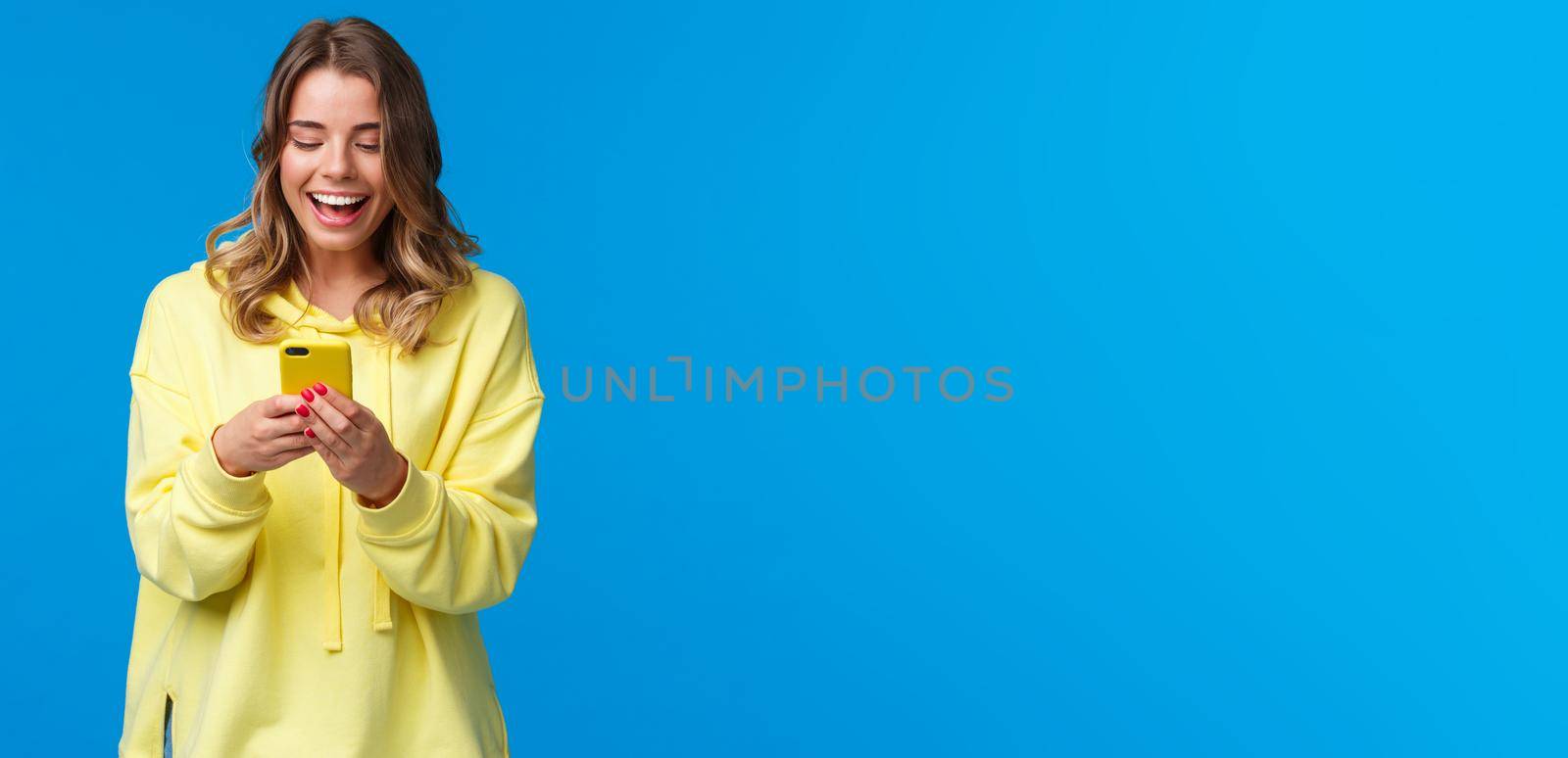 Technology and people concept. Happy smiling blond girl using mobile phone in yellow case, messaging look at smartphone display with pleased expression, send funny meme, blue background by Benzoix