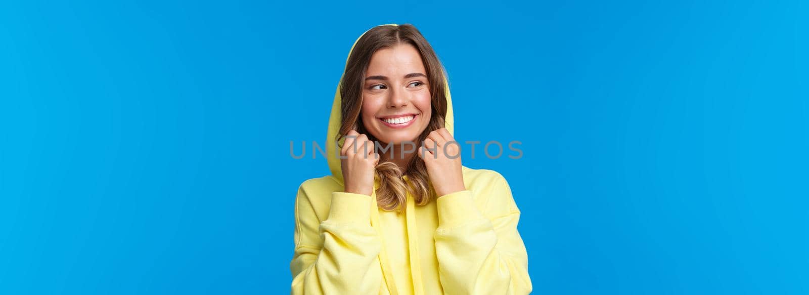 Close-up portrait beautiful charming blond girl put on hood of sweatshirt and smiling, look away, feel chilly as walking on spring day, standing carefree over blue background. Copy space