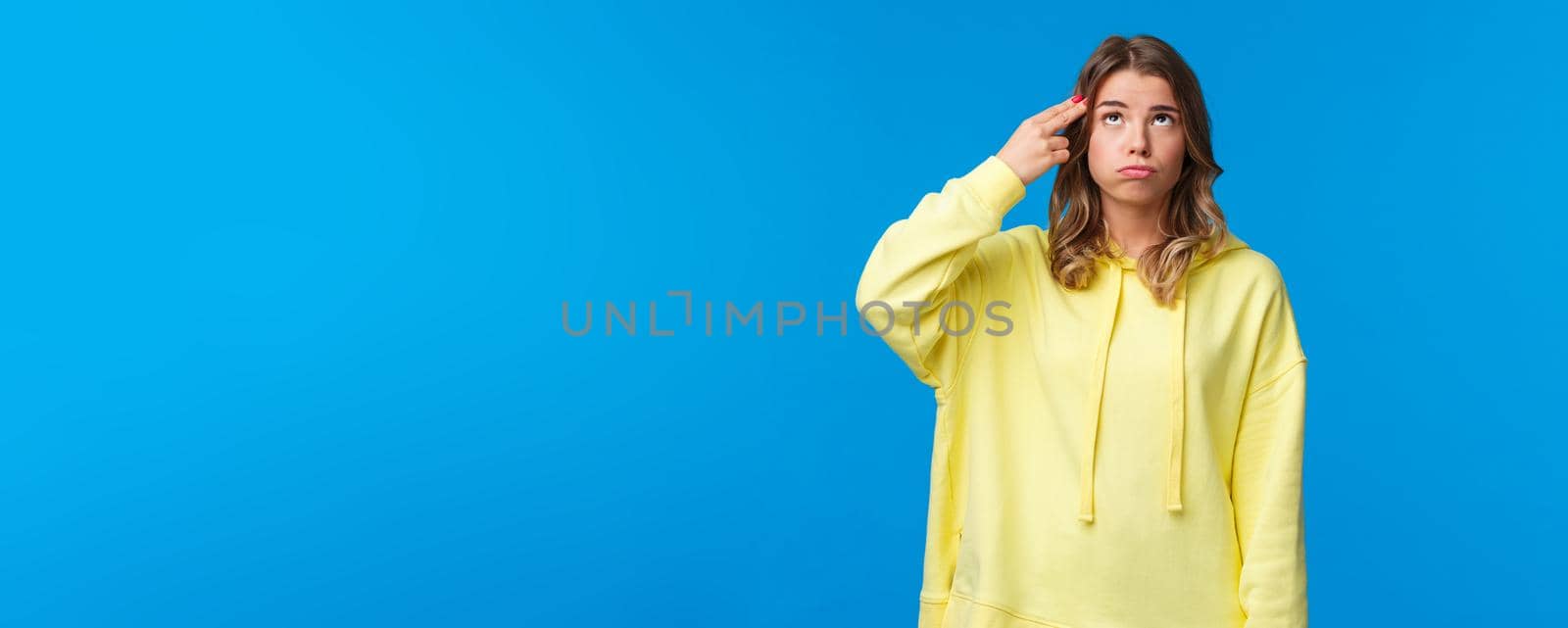 Gosh just kill me. Bored and pressured, stressed young blonde girl in yellow hoodie, make pistol gesture near head, shooting from gun and sighing as feel bothered and troubled, blue background by Benzoix