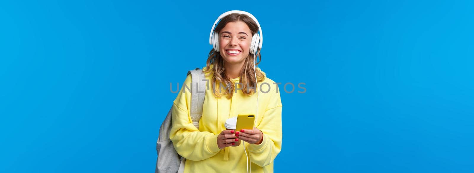 Cheerful pretty young female student with backpack, listening favorite playlist in headphones, laughing and smiling at camera carefree, hold mobile phone, stand blue background.