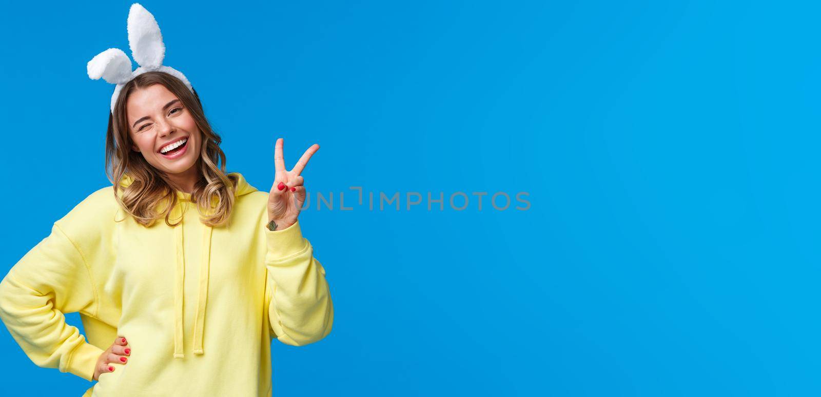 Holidays, traditions and celebration concept. Lovely and tender blond girl in yellow hoodie, tilt head kawaii wink and smiling as wearing rabbit ears, show peace gesture for goodwill, happy easter by Benzoix