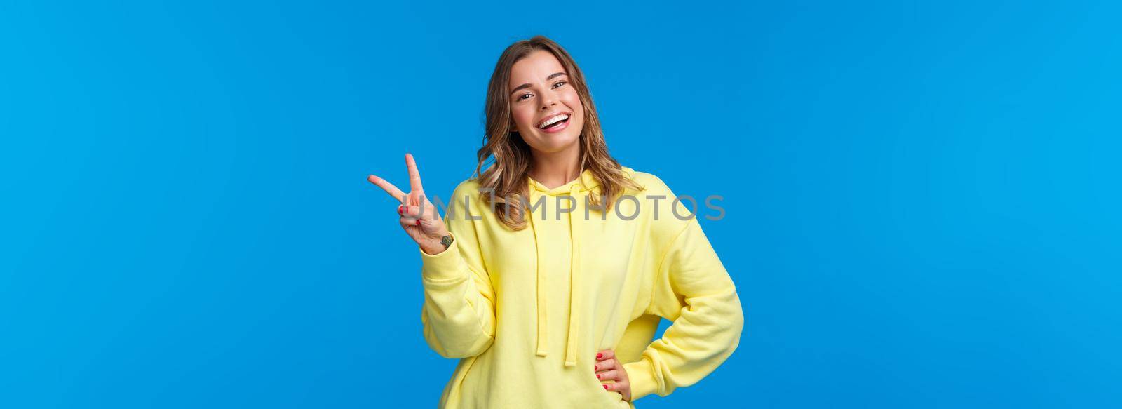 Staying bright sie. Carefree happy young silly blond girl in yellow hoodie sending positive vibes, show kawaii peace sign and smiling with white teeth, standing blue background.