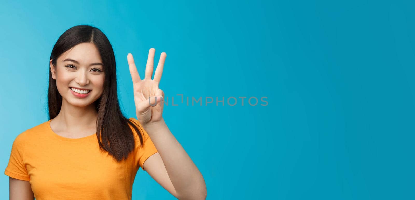 Friendly enthusiastic tender asian woman making reservation for three, number third, smiling broadly, stand blue background joyfully, posing near blue background in yellow t-shirt.