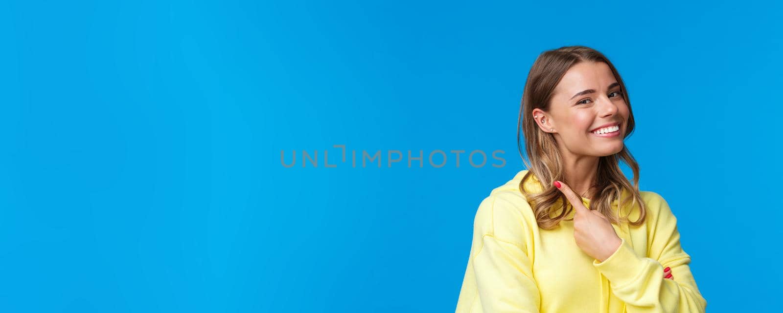 Close-up portrait of beautiful blond girl with short hair pointing at her piercing in ear, smiling proud and satisfied, change her look, standing blue background in yellow hoodie. Copy space