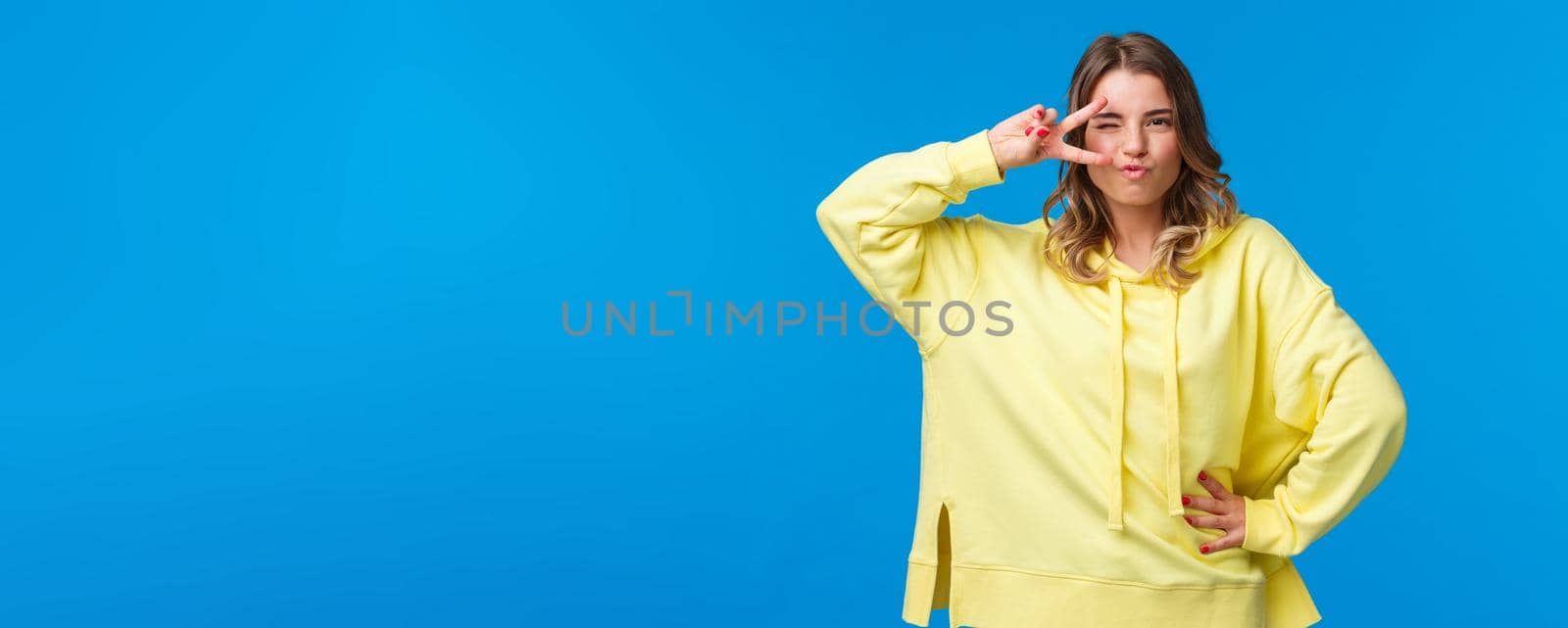 Portrait of sassy alluring blond caucasian woman dancing disco, show peace sign near eye and looking daring and sexy camera, standing in assured pose over blue background.