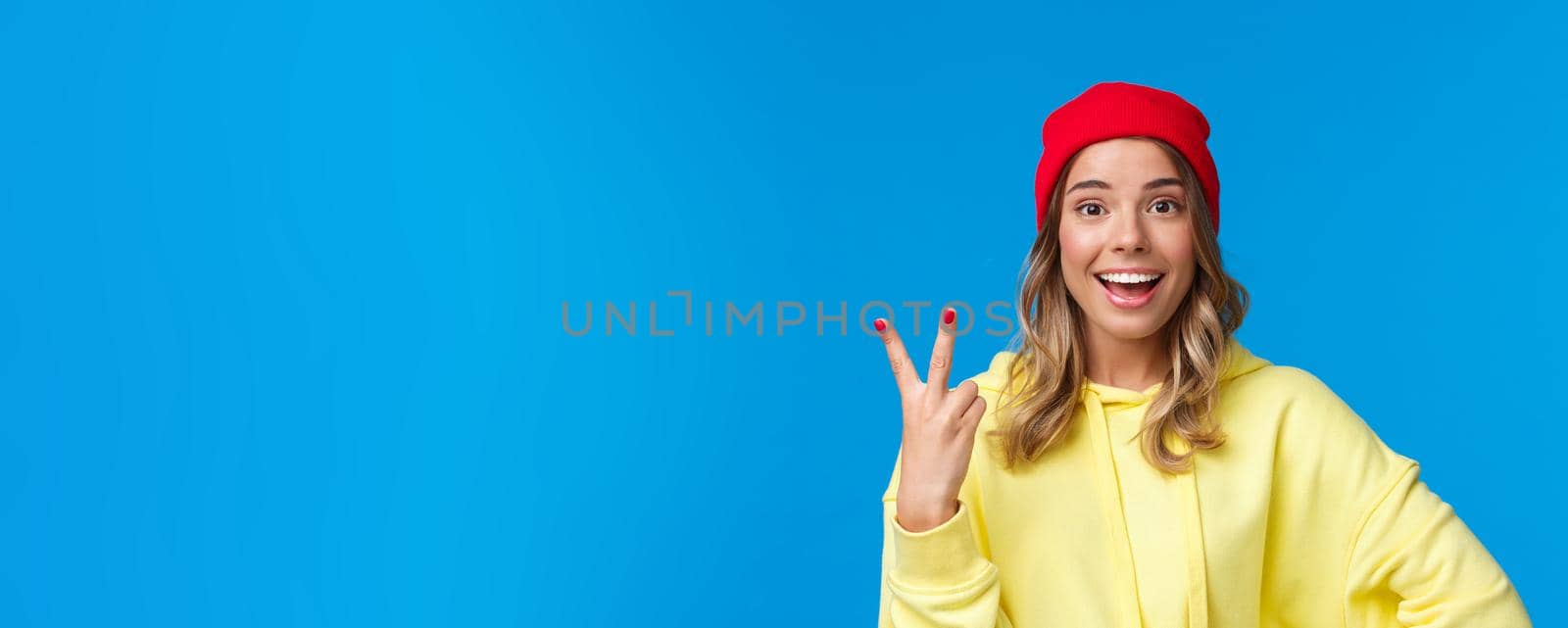 Cheerful good-looking caucasian blond in red beanie smiling happy, showing two fingers, making order, being second, standing blue background upbeat in red beanie and hoodie.