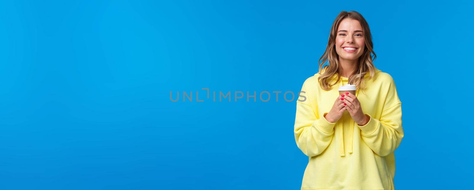 Girl enjoying tasty cup of morning coffee from favorite cafe. Cheerful lovely blond woman in yellow hoodie smiling white teeth and holding paper cup of take-away tea, stand blue background.