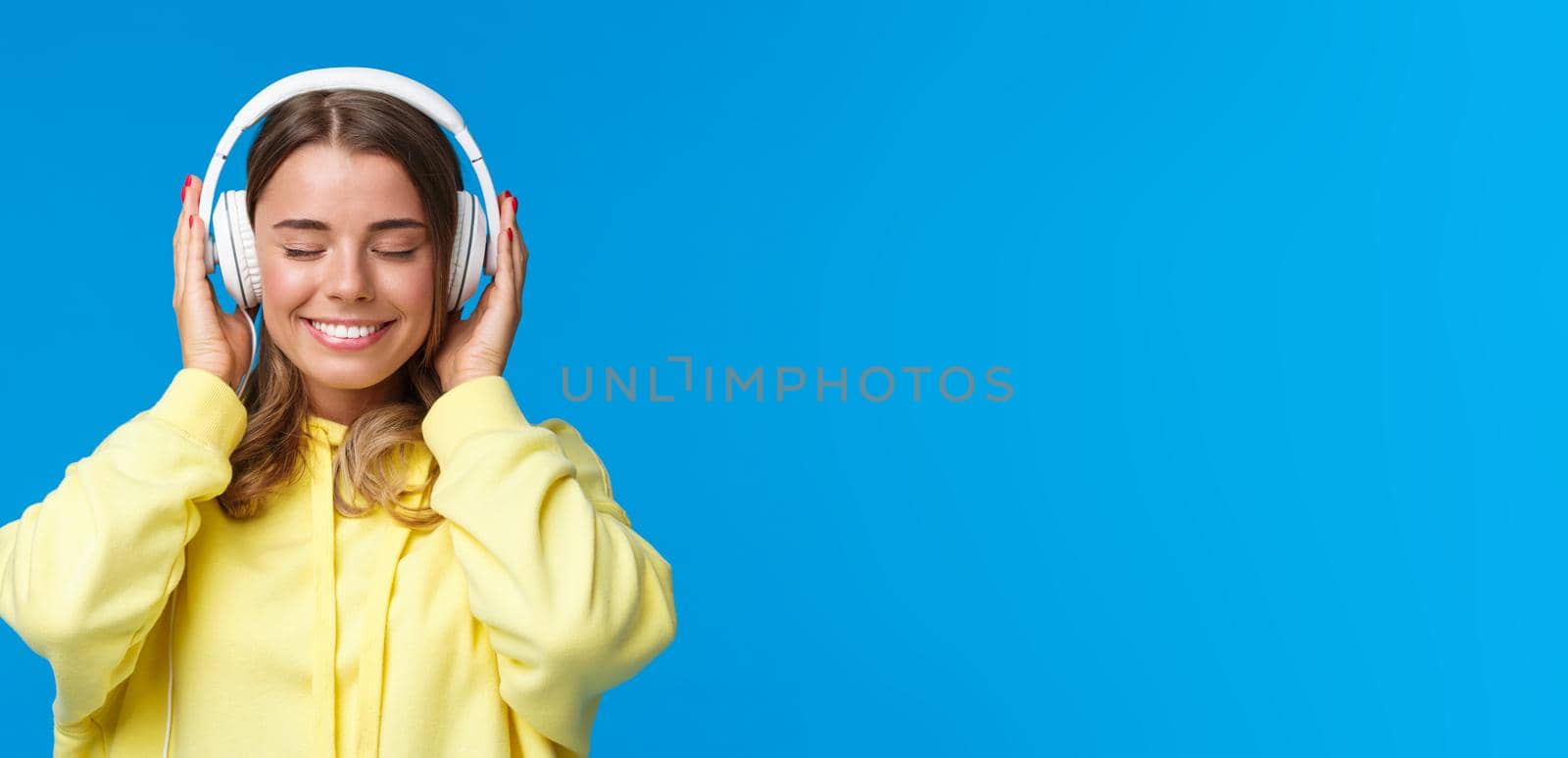 Music, lifestyle and youth concept. Close-up portrait of dreamy happy and relaxed young blond european girl close eyes and enjoying fine sound of song in headphones, dreaming, blue background.