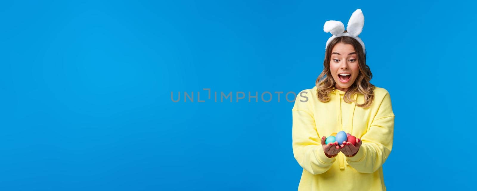 Surprised emotive cute blond caucasian girl playing holiday game, found Easter eggs look amused, wearing rabbit ears as celebrating religious holiday on sunday, blue background by Benzoix