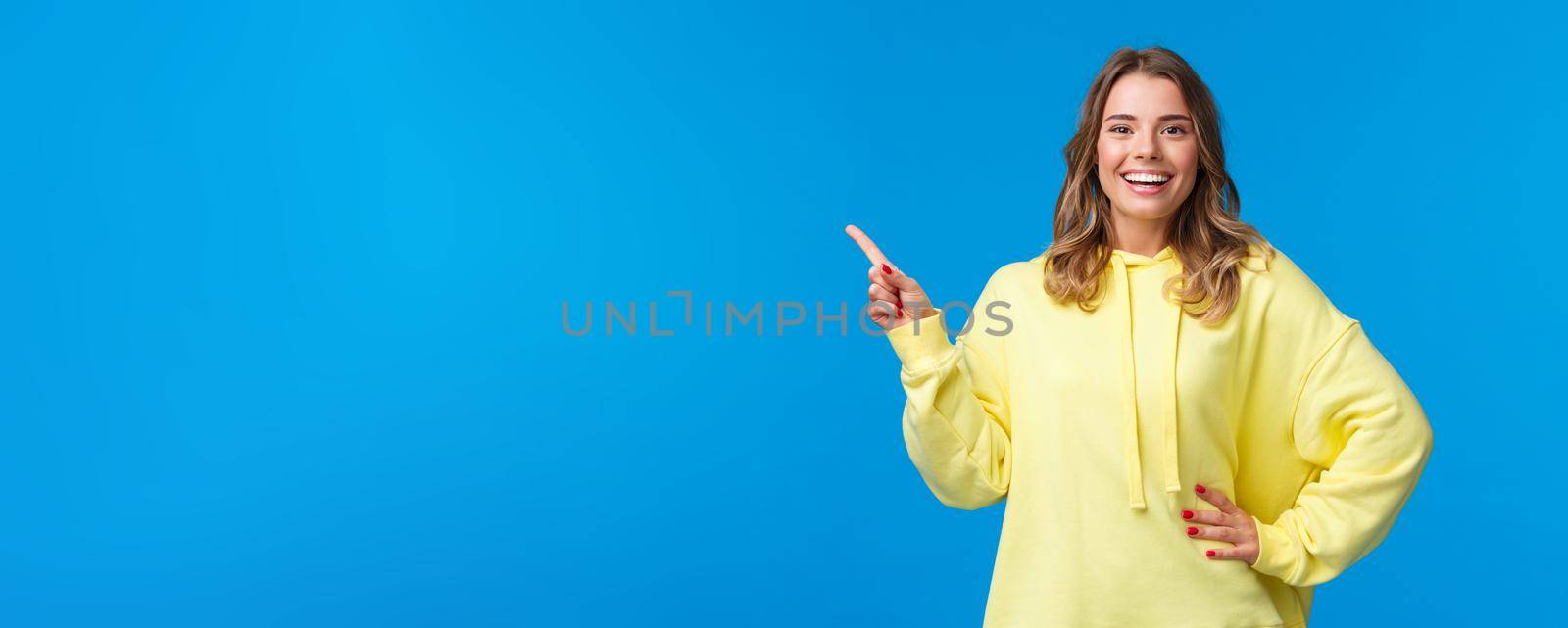 Take a look. Upbeat good-looking blond girl pointing finger left, recommend promo, advertise product, look pleased and smiling camera, explain project, standing blue background.