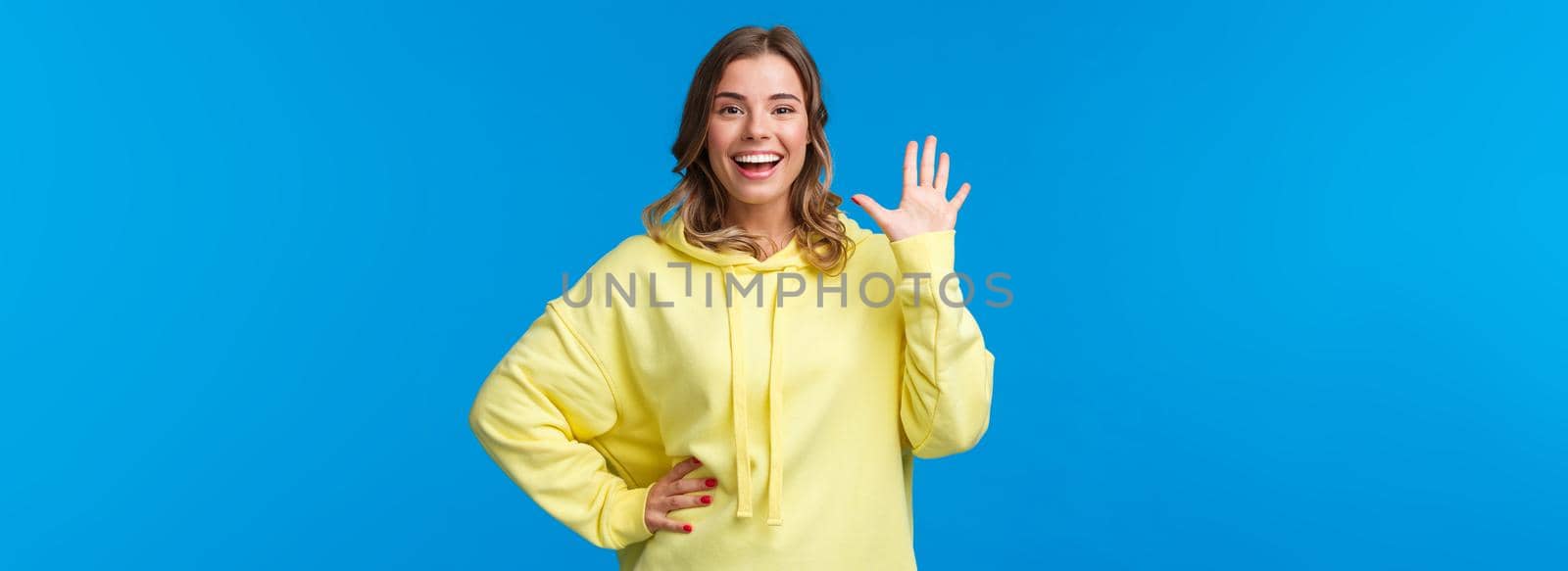 Cheerful friendly outgoing blond girl waving hand, informal greeting concept, smiling white teeth saying hi, welcoming new members to company, see friend, stand blue background.