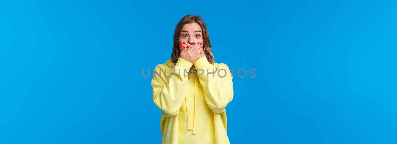 Shocked and speechless impressed blond woman close mouth with hands and stare astonished, hear shook rumour, gossiping make astounded face, standing blue background.