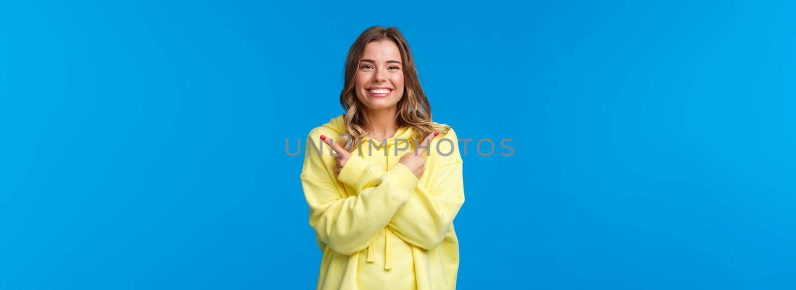 Confident pleasant, friendly-looking caucasian female showing two variants, give opportunity to choose what best, pointing fingers sideways and smiling camera, blue background.