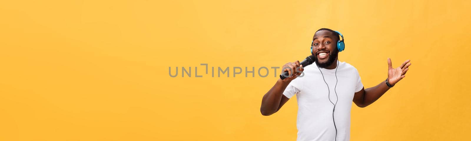 Portrait of cheerful positive chic handsome african man holding microphone and having headphones on head listening music singing song enjoying weekend vacation isolated on yellow background by Benzoix