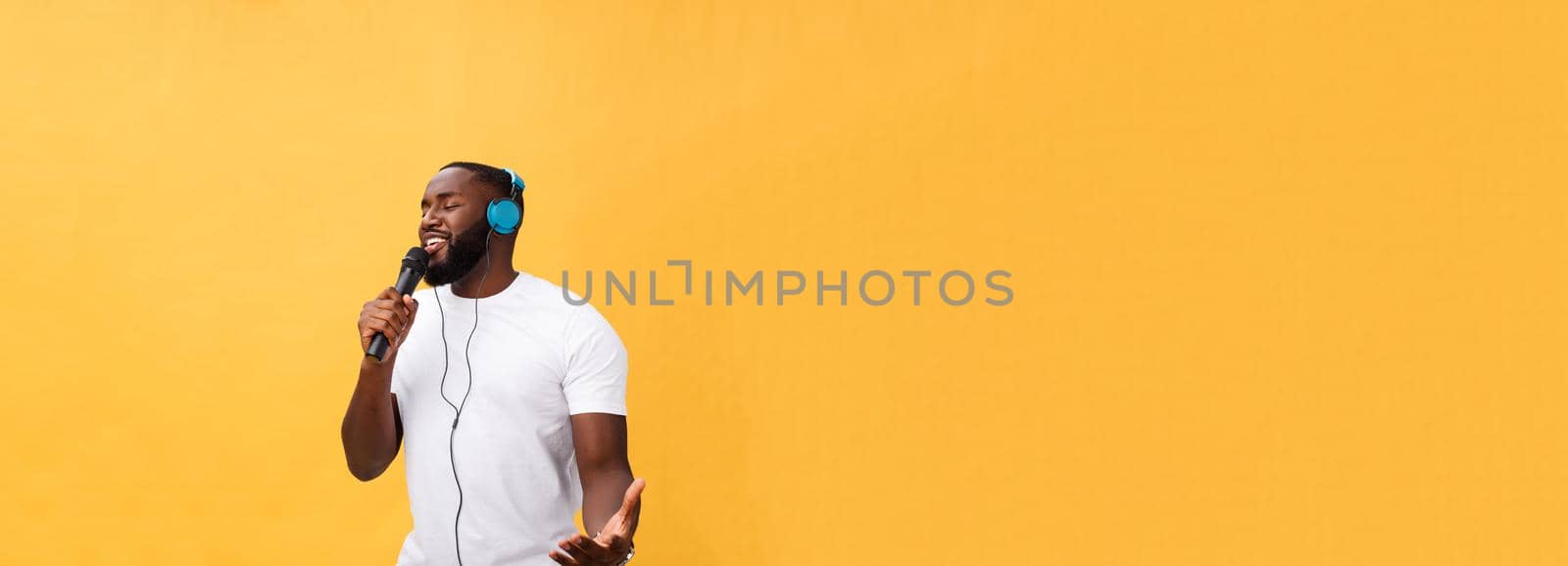 Portrait of cheerful positive chic. handsome african man holding microphone and having headphones on head listening music singing song enjoying weekend vacation isolated on yellow background.